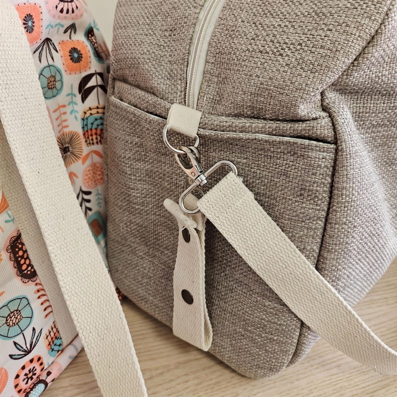 May include: A textured beige duffel bag with a cream-colored fabric shoulder strap attached by a silver metal swivel hook. Beside it sits a bag featuring a white fabric with a repeating pattern of stylized orange, teal, and black floral motifs.