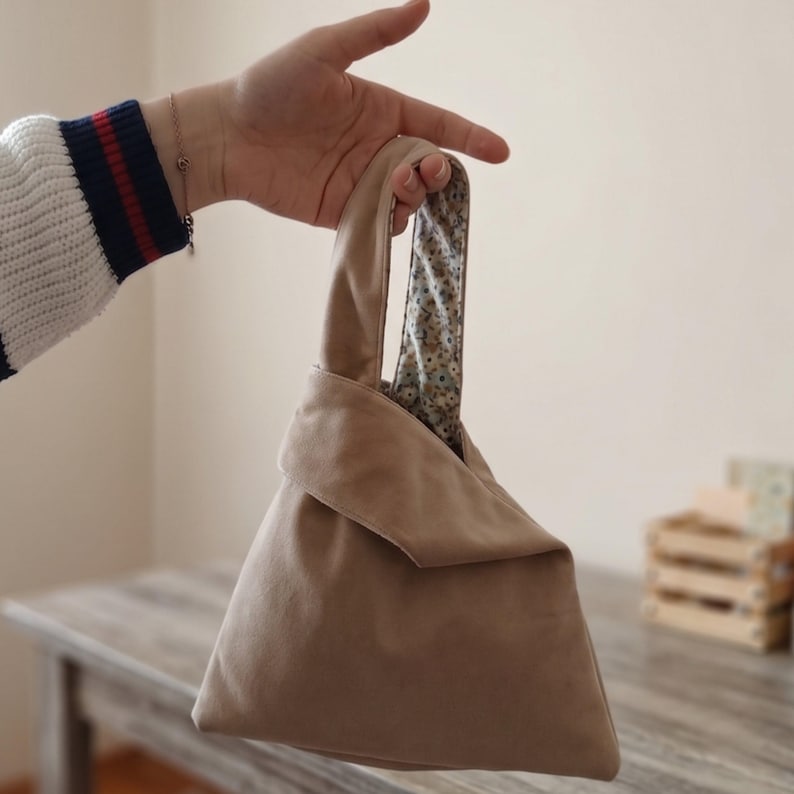 Peut inclure: Un sac &agrave; main en tissu beige avec une silhouette triangulaire et une poign&eacute;e unique. L'ext&eacute;rieur est d'une couleur marron clair unie, tandis que la doublure int&eacute;rieure pr&eacute;sente un motif floral &agrave; petite &eacute;chelle avec des fleurs bleues, jaunes et vertes.