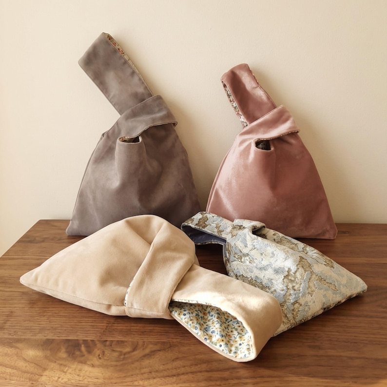 Peut inclure: Quatre sacs &agrave; n&oelig;ud de style japonais en velours et tissu &agrave; motifs. Un sac est gris, un est rose poudr&eacute;, un est beige et un pr&eacute;sente un motif floral textur&eacute; bleu et dor&eacute;. Chaque sac poss&egrave;de une anse unique qui passe dans l'autre.