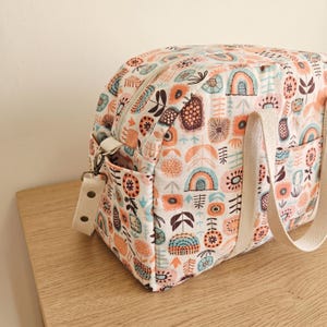 May include: A quilted fabric bag featuring a repeating pattern of stylized flowers, rainbows, and geometric shapes in shades of orange, teal, and dark brown. The bag includes a top zipper closure, side pockets, and cream-colored webbing straps.