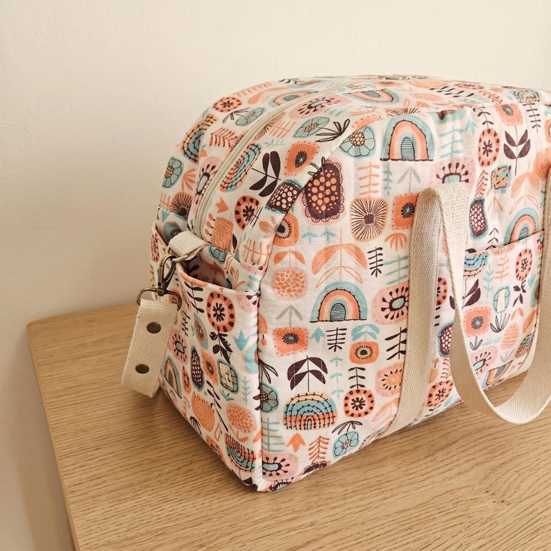 May include: A quilted fabric bag featuring a repeating pattern of stylized flowers, rainbows, and geometric shapes in shades of orange, teal, and dark brown. The bag includes a top zipper closure, side pockets, and cream-colored webbing straps.