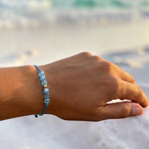Puede incluir: Una pulsera de cordón azul que presenta una serie de pequeñas cuentas rectangulares de color azul claro y una cuenta metálica de tono plateado en el centro. La pulsera se asegura con un cordón azul fino y ajustable alrededor de la muñeca.