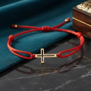 Puede incluir: Una pulsera de cordón rojo con un dije central de cruz en tono dorado con diseño de marco abierto. La pulsera incluye nudos deslizantes ajustables y pequeñas cuentas en tono dorado en los extremos del cordón, creando una pieza sencilla.