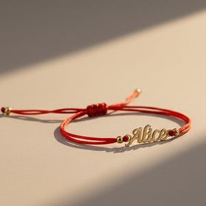 Puede incluir: Una pulsera de cordón rojo con una placa de metal dorado que lleva el nombre Alice en letra cursiva. La pulsera incluye pequeñas cuentas doradas en los extremos y un cierre de nudo corredizo para ajustar el tamaño a la muñeca.