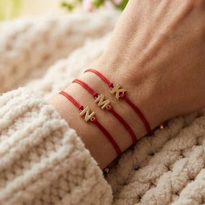 Peut inclure: Trois bracelets fins en cordon rouge, chacun orné d'une breloque en métal doré en forme de lettre. Les breloques affichent les lettres K, M et N. Les bracelets sont portés superposés, mettant en valeur le design minimaliste et la couleur rouge.