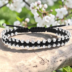 Op de afbeelding: Een zwart koordarmband met twee parallelle rijen kleine, gepolijste zilverkleurige metalen kralen. De armband is gemaakt met een geweven macramé-knoopmotief en bevat een verstelbaar schuifsluitingsmechanisme aan de achterzijde.