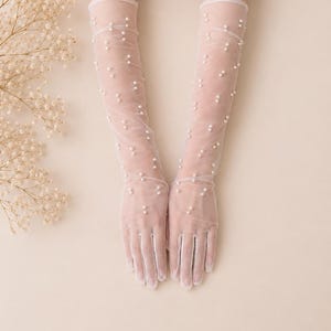 Könnte beinhalten: Ein Paar lange, transparente Handschuhe aus weißem Netzstoff. Die Handschuhe sind mit kleinen, weißen Kunstperlen verziert, die über die gesamte Länge der Arme und Hände verteilt sind und von den Fingerspitzen bis zum Unterarm reichen.