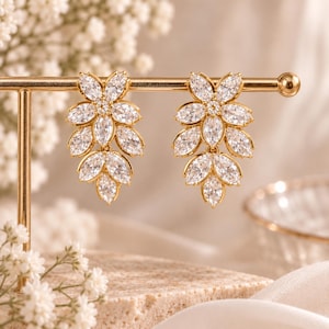 Könnte beinhalten: Ein Paar goldfarbene Ohrringe mit einer Anordnung aus klaren, marquise-geschliffenen Zirkonia-Steinen in Blattform. Jeder Ohrring besteht aus mehreren facettierten weißen Steinen, die in einem metallischen Rahmen gefasst und an einer Stange befestigt sind.