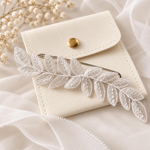 Könnte beinhalten: Eine silberfarbene Haarspange mit einem länglichen, weiß bestickten Blattmotiv. Das Accessoire liegt auf einem kleinen, cremefarbenen quadratischen Beutel mit einem goldfarbenen Druckknopfverschluss, der die detaillierte Stickerei der Blätter zeigt.