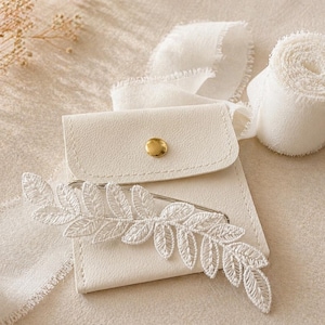 May include: A white, leaf-shaped lace brooch with a silver pin, on a small, off-white leather pouch with a gold button. A roll of white ribbon and a piece of white fabric are also in the image.