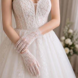 Peut inclure: Une paire de gants en dentelle blanche avec un motif floral délicat et des bords festonnés, portés avec une robe blanche sans bretelles dotée d'un décolleté en cœur et d'une broderie en dentelle complexe sur le corsage et la jupe.