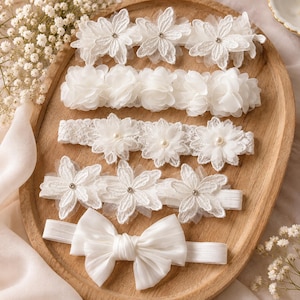 Può includere: Un set di cinque fasce per capelli bianche disposte su un vassoio di legno. La collezione comprende tre fasce con applicazioni in pizzo floreale decorate con piccoli strass o perle, una fascia con petali in tessuto e una fascia con un grande fiocco in raso.