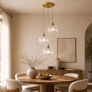 May include: Three pendant lights with clear, ribbed glass shades hang from a brass ceiling fixture. The lights are suspended above a round wooden dining table with cream-colored chairs. The room has a neutral color palette with a warm, inviting atmosphere.