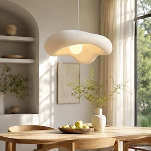 Peut inclure: Un luminaire suspendu de forme organique, de couleur crème, est suspendu au-dessus d'une table à manger en bois. La lumière a une surface douce et texturée et une ampoule chaude et brillante. La table est ornée d'un bol en bois de poires et d'un vase blanc avec de la verdure.