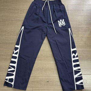 May include: Navy blue track pants with white accents and the word "AMIRI" printed vertically down the sides. The pants have a drawstring waist, white zippers, and a white logo on the left leg.