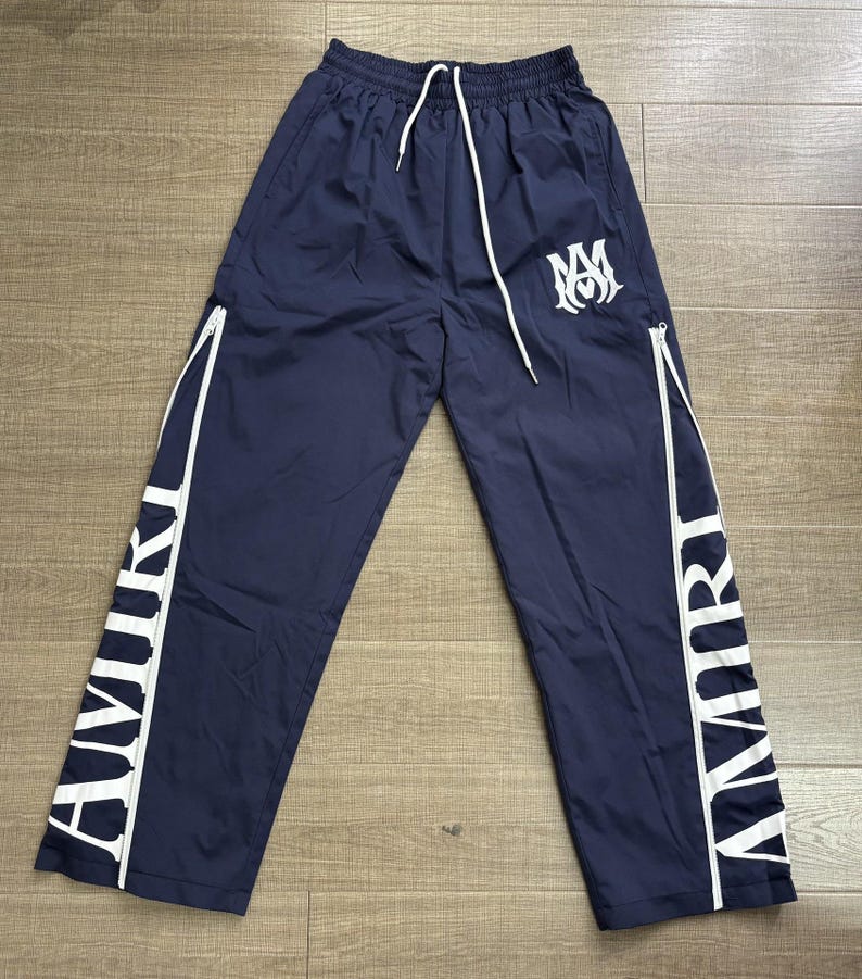 May include: Navy blue track pants with white accents and the word "AMIRI" printed vertically down the sides. The pants have a drawstring waist, white zippers, and a white logo on the left leg.