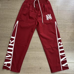 May include: Maroon track pants with white accents and the word "AMIRI" printed vertically on the sides. The pants have a white drawstring, a white logo, and white zippers. The pants are made of a smooth, lightweight material.