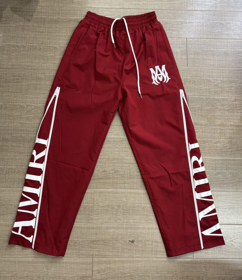 May include: Maroon track pants with white accents and the word "AMIRI" printed vertically on the sides. The pants have a white drawstring, a white logo, and white zippers. The pants are made of a smooth, lightweight material.
