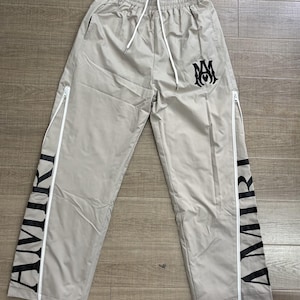 May include: Light grey track pants with white zippers and drawstring. The word "AMIRI" is printed vertically in black on the sides. A black logo is on the upper right leg.