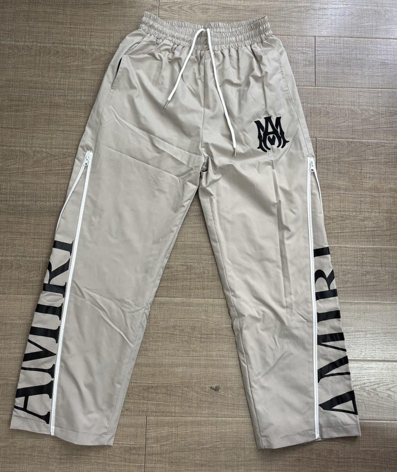 May include: Light grey track pants with white zippers and drawstring. The word "AMIRI" is printed vertically in black on the sides. A black logo is on the upper right leg.
