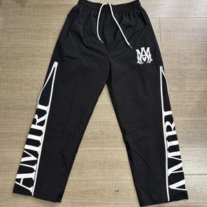 May include: Black athletic pants with white accents and the word "AMIRI" printed vertically on the sides. The pants have a drawstring waist, white zippers, and a white logo on the left leg.