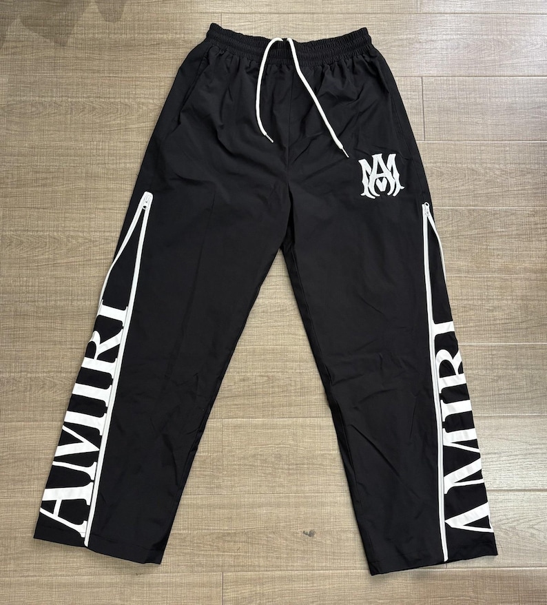 May include: Black athletic pants with white accents and the word "AMIRI" printed vertically on the sides. The pants have a drawstring waist, white zippers, and a white logo on the left leg.
