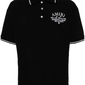 Puede incluir: Polo negro con ribetes blancos en el cuello y las mangas. La camisa presenta el nombre de la marca "AMIRI" y el texto "IMAGINATION AND DREAMS ARTS AND CRAFTS" en letras blancas.