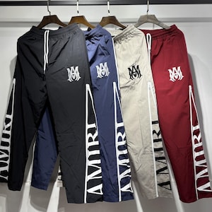 May include: A collection of track pants in black, navy blue, beige, and burgundy. Each pair features a white zipper detail, the word "AMIRI" printed vertically on the leg, and a logo on the upper left leg.