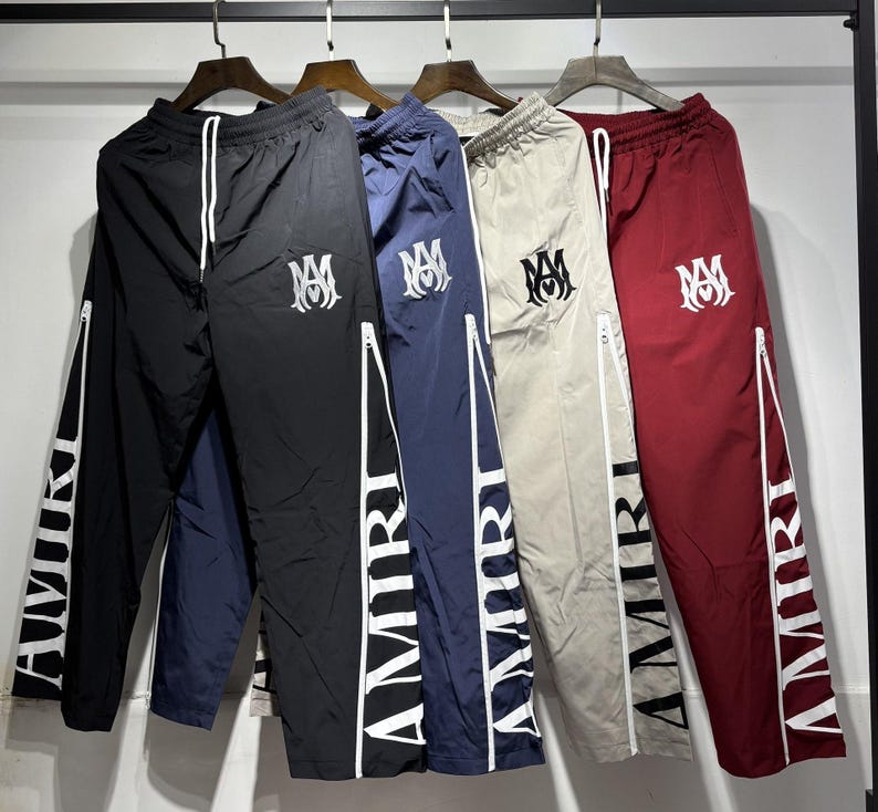 May include: A collection of track pants in black, navy blue, beige, and burgundy. Each pair features a white zipper detail, the word "AMIRI" printed vertically on the leg, and a logo on the upper left leg.