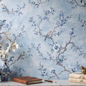May include: A light blue floral wallpaper with branches, blue flowers, and birds. Two blue and white vases with flowers are in front of the wallpaper. A brown leather journal, pen, and books are on a white surface.
