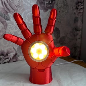 May include: A metallic red robotic hand lamp featuring a circular palm light with six illuminated white LED bulbs. The hand is positioned in an open palm gesture, with segmented fingers and a smooth, glossy finish, connected to a white power cord.