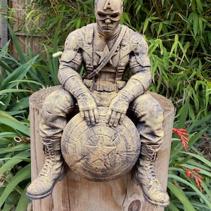 May include: A gold-toned, textured figurine of Captain America sitting on a wooden post. The figure wears a detailed tactical suit with a star emblem on the chest and a helmet with an 'A' on the forehead. Hands rest on a circular shield.
