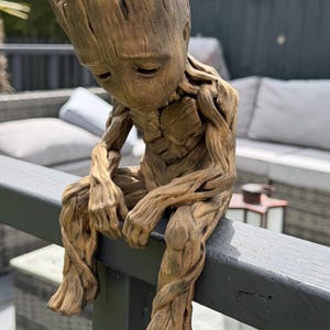 May include: A brown, tree-like humanoid figurine with a textured, bark-like surface. The figure has a large head, downward-gazing eyes, and slender limbs, depicted in a seated position with its legs dangling and arms resting on its knees.