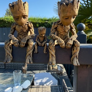 May include: Three brown, textured tree-like figures with humanoid features, including two larger figures and one smaller figure positioned between them, all sitting with their heads bowed and arms resting on their knees in a contemplative pose.