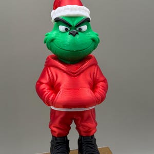 May include: A green character figurine with a mischievous expression, wearing a red Santa hat with a white brim, a red hooded sweatshirt, red trousers, and black high-top trainers with white soles. The figure stands upright with hands in pockets.