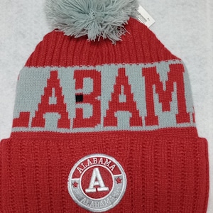May include: Red knit beanie with a gray pom pom on top. The beanie has a gray band with the word "BAMA" in red letters. The beanie also has a gray patch with the word "ALABAMA" and a white letter "A" in the center.