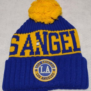 May include: Blue and yellow knit beanie with a pom pom on top. The beanie has the words "Los Angeles" and "LA" embroidered on it.