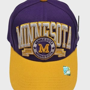 May include: A purple and gold baseball cap with "MINNESOTA" embroidered across the front in white. The cap features a circular emblem with "MINNESOTA" and the letter "M". The brim is gold, and the top button is also gold.