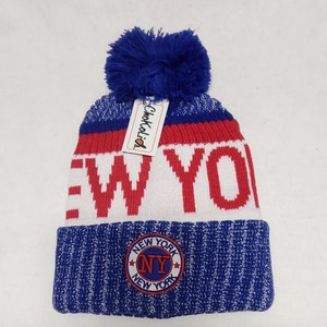 May include: A blue, red, and white knit beanie with a blue pom-pom. The word "NEW YORK" is displayed in red block letters on the front. A circular patch with "NEW YORK" and "NY" is on the brim. A tag with the brand name "ChokoKali" is attached.