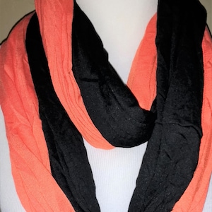 May include: A coral and black infinity scarf. The scarf is draped around a white mannequin. The scarf is made of a soft, lightweight fabric. The coral and black colors are arranged in alternating stripes.
