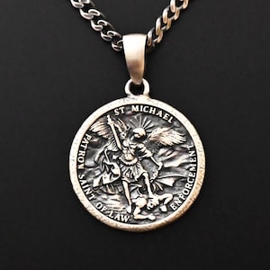 May include: Silver-toned pendant necklace featuring a round medallion with the image of St. Michael. The medallion has the words "ST MICHAEL", "PATRON SAINT OF LAW ENFORCEMENT". The chain is a curb link style. The background is black.