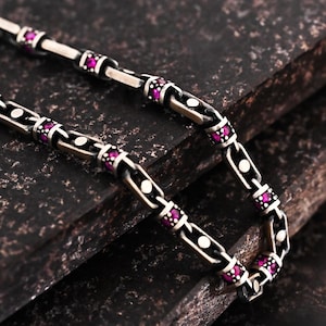 May include: A silver chain necklace featuring rectangular links with oxidized detailing. Each link is accented with small, round, vibrant pink gemstones and a single white circular inlay, creating a repeating pattern along the entire length.