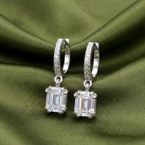 May include: A pair of silver-toned hoop earrings featuring a row of small clear stones along the hoop and a single emerald-cut clear stone pendant hanging from each hoop. The earrings have a polished metallic finish and a secure hinged closure.