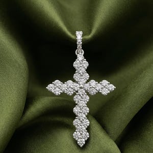 May include: A silver-toned cross pendant featuring a cluster of round, clear, sparkling stones arranged in a floral-like pattern. The pendant includes a small, stone-encrusted bail at the top for attachment to a chain, showcasing a bright, reflective metallic finish throughout the entire piece.