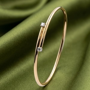 May include: A thin, polished gold bangle bracelet featuring an open-ended design. Two small, round, clear faceted stones are set at the ends of the gold band, positioned to face each other with a slight gap between them, creating a sleek, refined look.