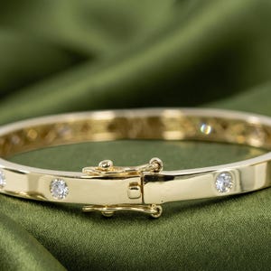 Könnte beinhalten: Ein aufklappbares Armband aus Gold mit einer Reihe von runden, klaren, facettierten Steinen, die bündig in das polierte Metallband eingelassen sind. Das Armband verfügt über einen sicheren, integrierten Kastenschloss-Mechanismus mit Sicherheitsbügel.