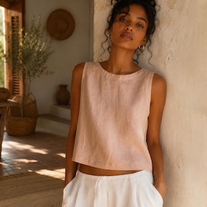May include: A sleeveless, light pink linen top with a round neckline. The top is paired with white linen pants. The model is leaning against a light-colored wall in a sunlit room. The overall aesthetic is casual and summery.