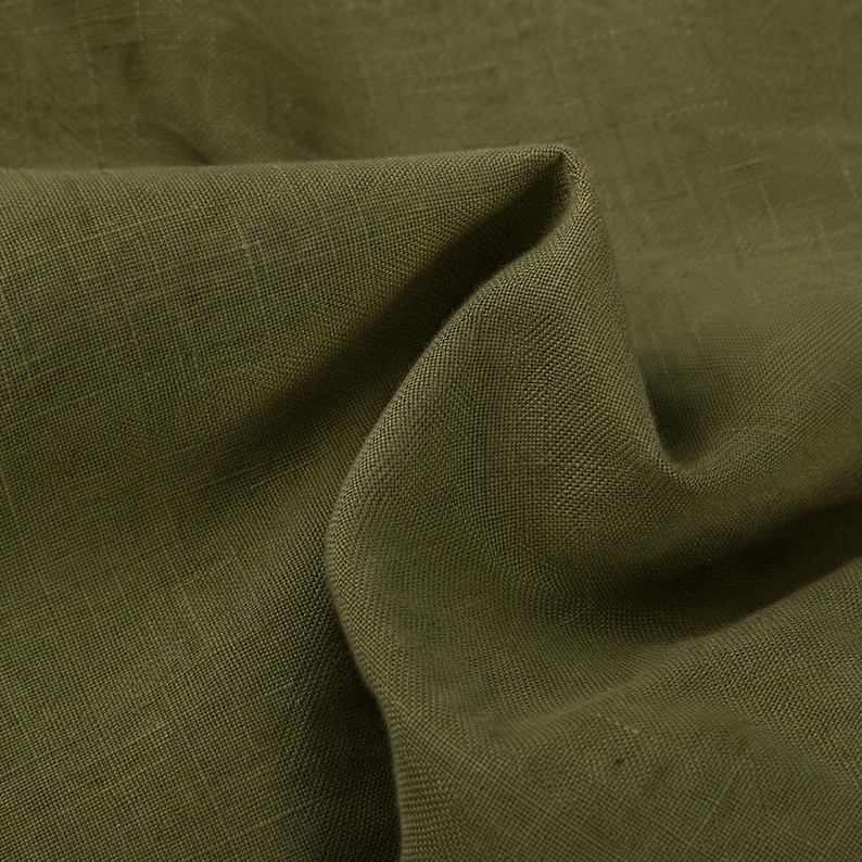 May include: A piece of olive green fabric with a visible woven texture. The material is folded to create soft, deep shadows and highlights across the surface, showcasing the natural weave pattern and the consistent, earthy tone of the textile.