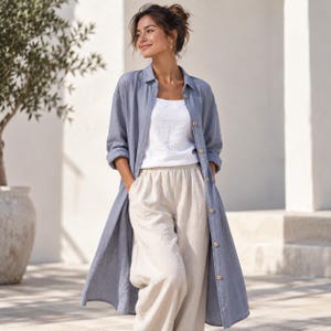 May include: A long-sleeved, button-down shirt in a muted blue color, worn open over a plain white tank top and loose-fitting beige trousers. The shirt features a classic collar and light-colored buttons, creating a layered outfit with a relaxed fit.