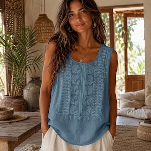 May include: A sleeveless blue top featuring white embroidered floral and geometric patterns on the front panel, a round neckline, and a relaxed fit. The garment is paired with light-coloured trousers, creating a simple and cohesive look.
